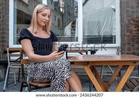 In this image, we see a serene young woman with blonde hair seated at a sidewalk cafe, deeply engaged with her smartphone. She wears a fashionable black blouse paired with a zebra-striped skirt