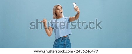 Lifestyle. Girl recording video showing followers awesome scene attending cool event holding smartphone looking impressed and surprised at device screen pointing backwards indicating right over blue