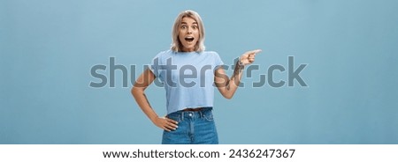 Studio shot of amazed impressed good-looking sportswoman in trendy t-shirt with tattoos opening mouth with enthusiasm from surprise holding hand on waist and pointing left over blue background