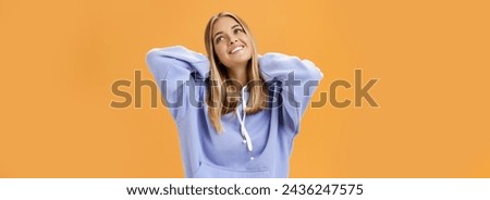 Young pleased and tender woman with tanned skin feeling cozy in warm in trendy hoodie touching back of neck smiling looking at upper right corner dreamy and delighted against orange background Royalty-Free Stock Photo #2436247575