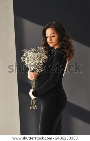 Studio portrait of beautiful pregnant woman in black dress holding a bouquet of gypsophila in her hands near pregnant belly. Pregnancy photosession. The concept of happy motherhood