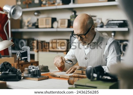 Business owner working in his shop and selling vintage items, he is checking an antique book using a magnifier