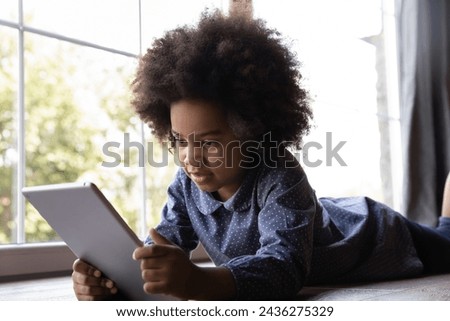 Cute African girl lying on floor indoor with digital tablet. Little 6s kid holds modern device play video game on internet. Generation z, secure usage, parental control app software concept