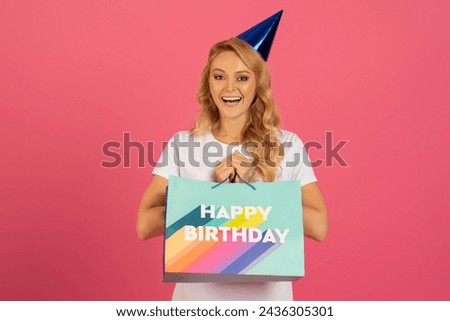 Festive Surprise. Happy blonde caucasian lady with party hat, joyfully holds her birthday gift against pink studio backdrop. Portrait shot of cheerful bday woman celebrating her special occasion