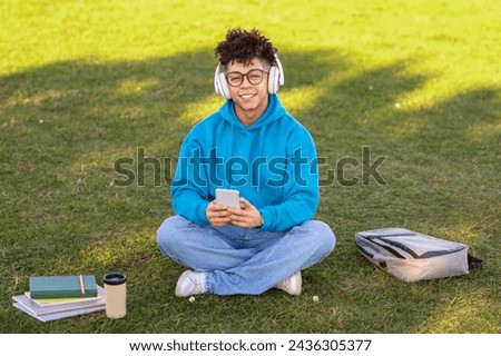 Smiling black student guy wearing headphones engages with mobile phone, listening to music and educational podcasts while learning outdoor, sitting with his backpack outside