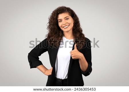 Cheerful young businesswoman with curly hair, in smart blazer, gives thumbs up sign while smiling confidently at the camera on grey studio backdrop