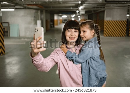 Happy family in casual clothes having video call using cell phone before exciting trip. Caucasian mother holding preschool daughter in hands making selfie with smartphone in underground parking lot.