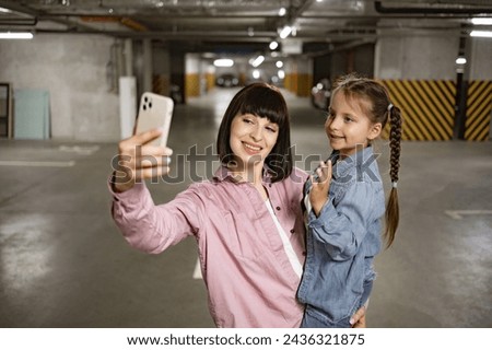 Happy family in casual clothes having video call using cell phone before exciting trip. Caucasian mother holding preschool daughter in hands making selfie with smartphone in underground parking lot.