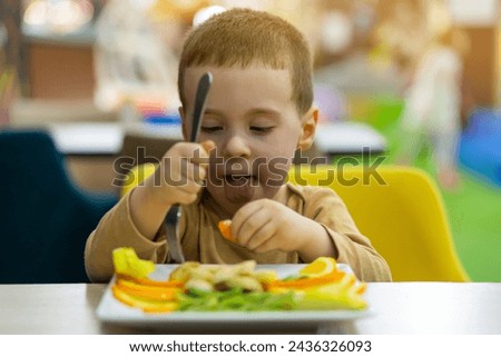 A funny cute three year old toddler boy eats fresh fruit while sitting at a table in a public place. Healthy food for children. Fruit plate. Focus on the face. Royalty-Free Stock Photo #2436326093