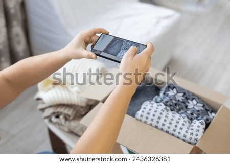 Clothing sweaters made from natural wool are folded into a box for recycling. A woman takes a photo of folded clothes on her phone and places an advertisement for sale on the Internet.
