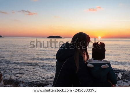 Silhouette of loving mother with small toddler walking on idyllic beach at sunset in coastal town Funtana, Istria, Croatia. Calm water surface of Adriatic Mediterranean Sea. Family vacation concept