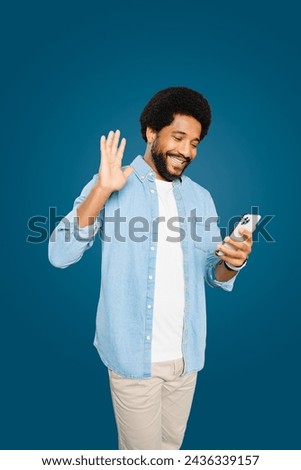 Handsome cheerful Brazilian young man with an afro hairstyle waving while holding his smartphone, portraying a friendly digital interaction or virtual greeting, involved video call or streaming online
