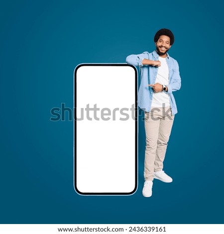 A young Brazilian man with a friendly smile stands beside a blank oversized smartphone, pointing at the potential place for advertising a new app or deal, male freelancer in denim shirt presenting Royalty-Free Stock Photo #2436339161