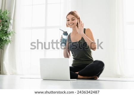 Happy young female athlete in sporty clothes sitting on fitness mat while talking on videocall on laptop. Online training, lesson of fitness. Social distancing concept Royalty-Free Stock Photo #2436342487