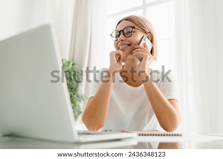 Happy cheerful caucasian young woman talking on the cellphone with customers clients working remotely on laptop. Freelance remote job, multitasking concept Royalty-Free Stock Photo #2436348123