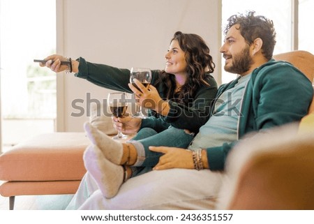 A smiling couple enjoys a television show together during a comfortable evening at home, each holding a glass of red wine - a picture of domestic bliss.
