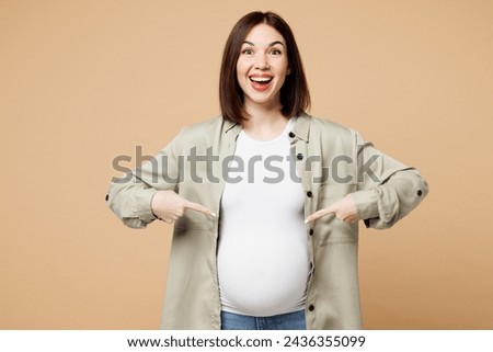 Young surprised fun pregnant expectant woman future mom wear grey shirt with belly stomach point index finger on tummy with baby isolated on plain beige background. Maternity family pregnancy concept