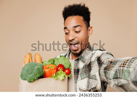 Young man wearing grey shirt hold paper bag for takeaway mock up with food products doing selfie shot on mobile cell phone isolated on plain beige background. Delivery service from shop or restaurant