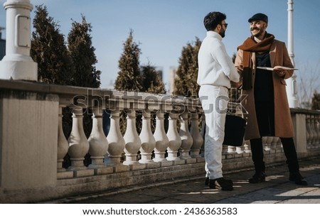 Smiling businessmen in stylish winter attire engage in a warm handshake outside, symbolizing successful collaboration or agreement. Royalty-Free Stock Photo #2436363583