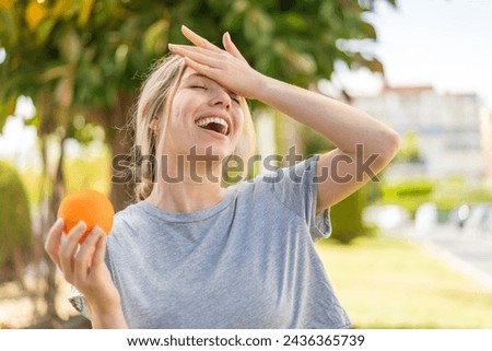 Young blonde woman holding an orange at outdoors has realized something and intending the solution