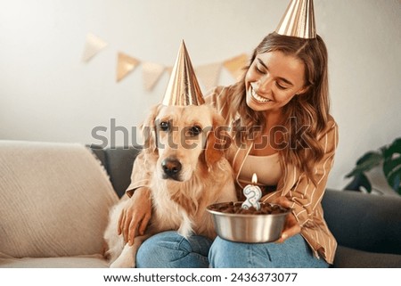 Young happy woman holding birthday cake, celebrating her pet dog's birthday, hugging and having fun with her pet while sitting in the living room at home.