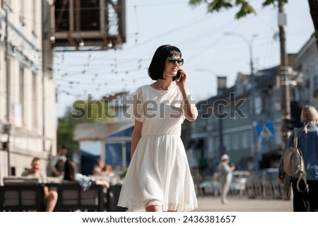 Romantic young brunette woman with short hair wears mini white dress and sunglasses, talking on cellphone, walking on a crowded city street in the summertime. Girl communicates by smartphone outdoors.