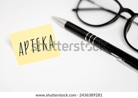 A yellow sticky note on a white background with the handwritten inscription "Apteka", next to it a black pen and glasses (selective focus) translation: pharmacy
