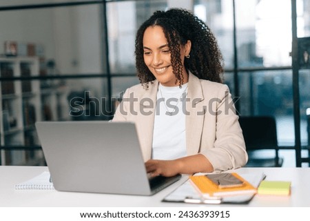 Amazing curly haired hispanic or brazilian young woman in stylish elegant clothes, office employee, small business owner, working on a laptop while sitting at her workplace in the modern office, smile Royalty-Free Stock Photo #2436393915