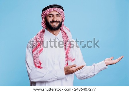 Muslim man in traditional robe confidently pointing to left side with hands studio portrait. Arab showcasing product advertisement and looking at camera with cheerful expression Royalty-Free Stock Photo #2436407297