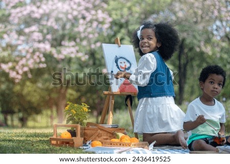 African little girl holds brush and paints picture on easel at park, very happy and smiling at camera, her younger brother sat next to her