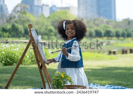 African little girl holds brush and paints picture on easel at park, very happy and smiling at camera, spend the holidays picnicking and painting