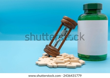 Medicine bottle with blank label, next to a stack of pills along with an hourglass isolated on blue background Daily dose concept with copy space Royalty-Free Stock Photo #2436431779