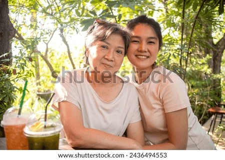 Asian Thai Chinese elder mother and daughter cuddling and take photo look at camera, drinking beverages at cafe garden, spend time together.