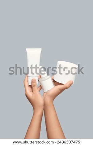 Closeup of beautiful woman hand holding white jar, white cream tube and white bottle of moisturizing serum. Isolated on grey background
