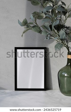 Blank poster mockup, black empty picture frame, green vase with tree branch on marble table, textured concrete wall background with soft natural sunlight shadows, elegant home interior decor.
