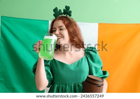 Young redhead woman with beer and barrel against flag of Ireland on green background. St. Patrick's Day celebration