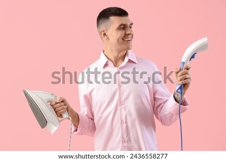 Happy young man with steamer and iron on pink background