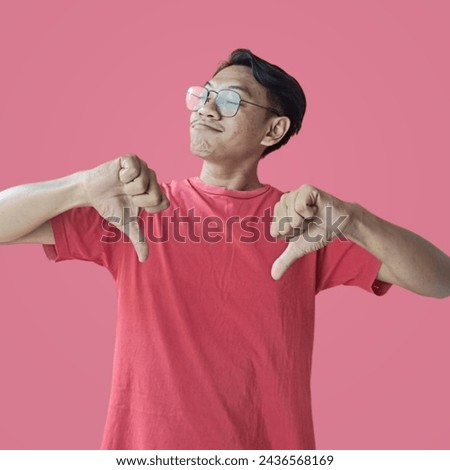 Portrait of a funny young Asian man doing mocking gesture with synical face and showing two thumbs down over pink background