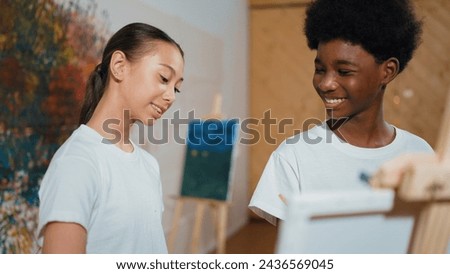 Cute girl looking canvas while boy painting picture in art lesson. Diverse highschool student standing at colorful stained wall while multicultural children attend in creative activity. Edification.