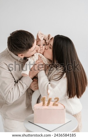 Dad and Mom kiss cute baby isolated on white wall. Father, mother hold hands funny little kid. Creative birthday cake with candle. Family celebrates six month old baby girl birthday party closeup.