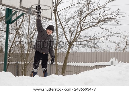 a boy stands on a snowdrift and holds on to a basketball hoop.