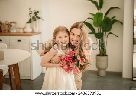 Mom and kid hugging, smiling. Happy family holiday. Cute little child girl greeting mother in kitchen. Beautiful daughter gives mother big bouquet of tulip flowers and makes surprise for Mothers Day.