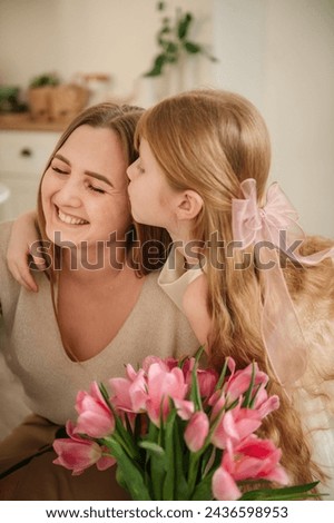 Daughter kisses and hugs mom. Happy family holiday. Cute little child girl greets smiling mother in kitchen. Beautiful kid gives mother big bouquet of tulip flowers and makes surprise for Mothers Day