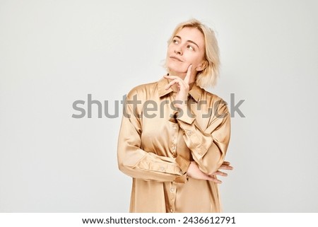 Thoughtful young woman in a stylish beige shirt posing with hand on chin against a white background.