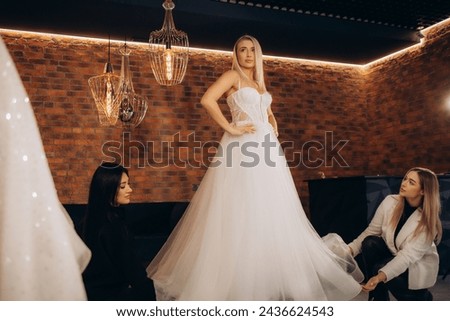 Beautiful bride getting dressed by her best friend in her wedding day and choosing a wedding dress in the shop and the shop assistant is helping her. High quality photo