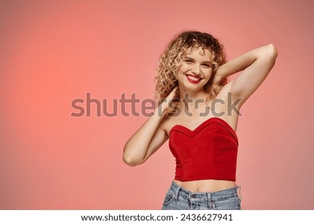 woman with red lips and curly hair posing in red top and smiling at camera on pastel backdrop