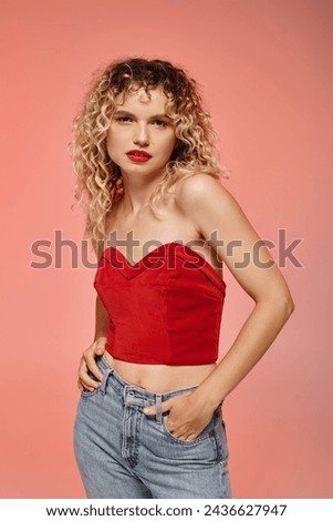 seductive woman with bold makeup posing in red top and looking at camera on pink backdrop