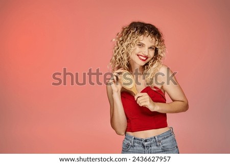 charming and curly woman in red trendy top posing with hair brush on yellow and pink backdrop