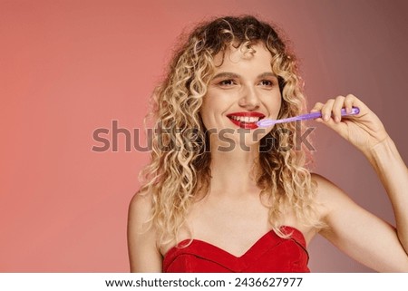 attractive curly woman with red lips and radiant smile cleaning teeth on pastel pink backdrop