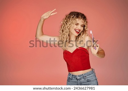 excited woman with wavy hair and red lips dancing with toothbrush on pink and yellow backdrop